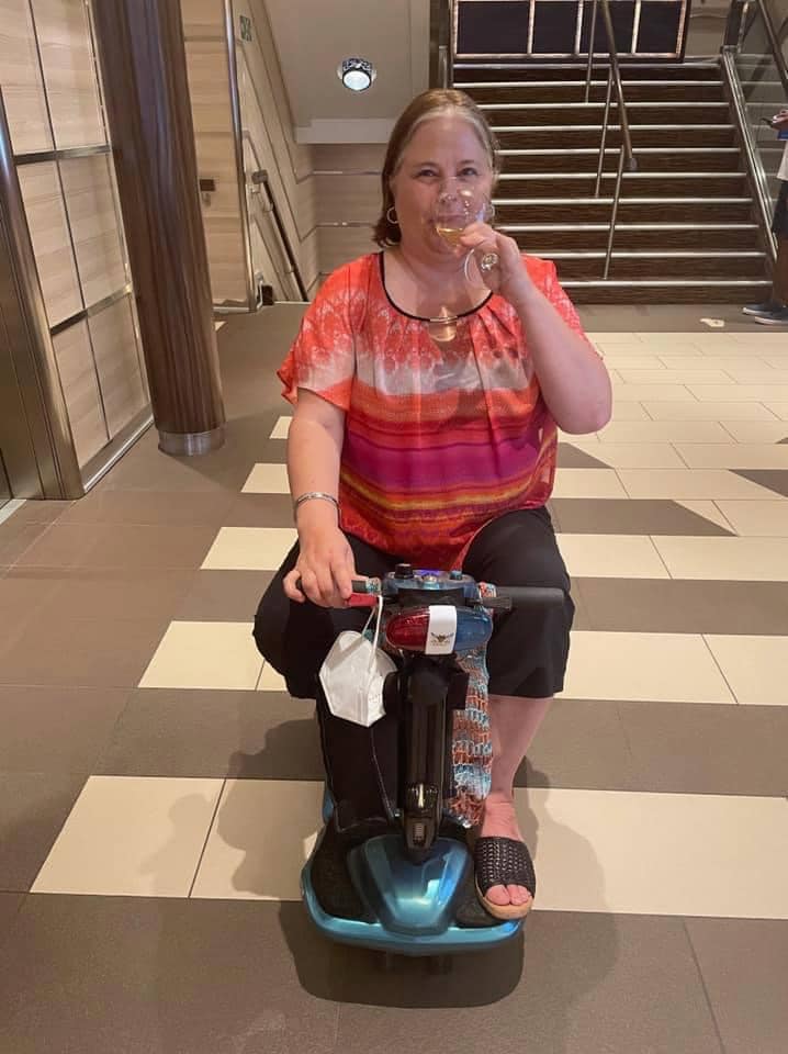 A woman sits atop a mobility scooter sipping a glass of wine. She is aboard a cruise ship